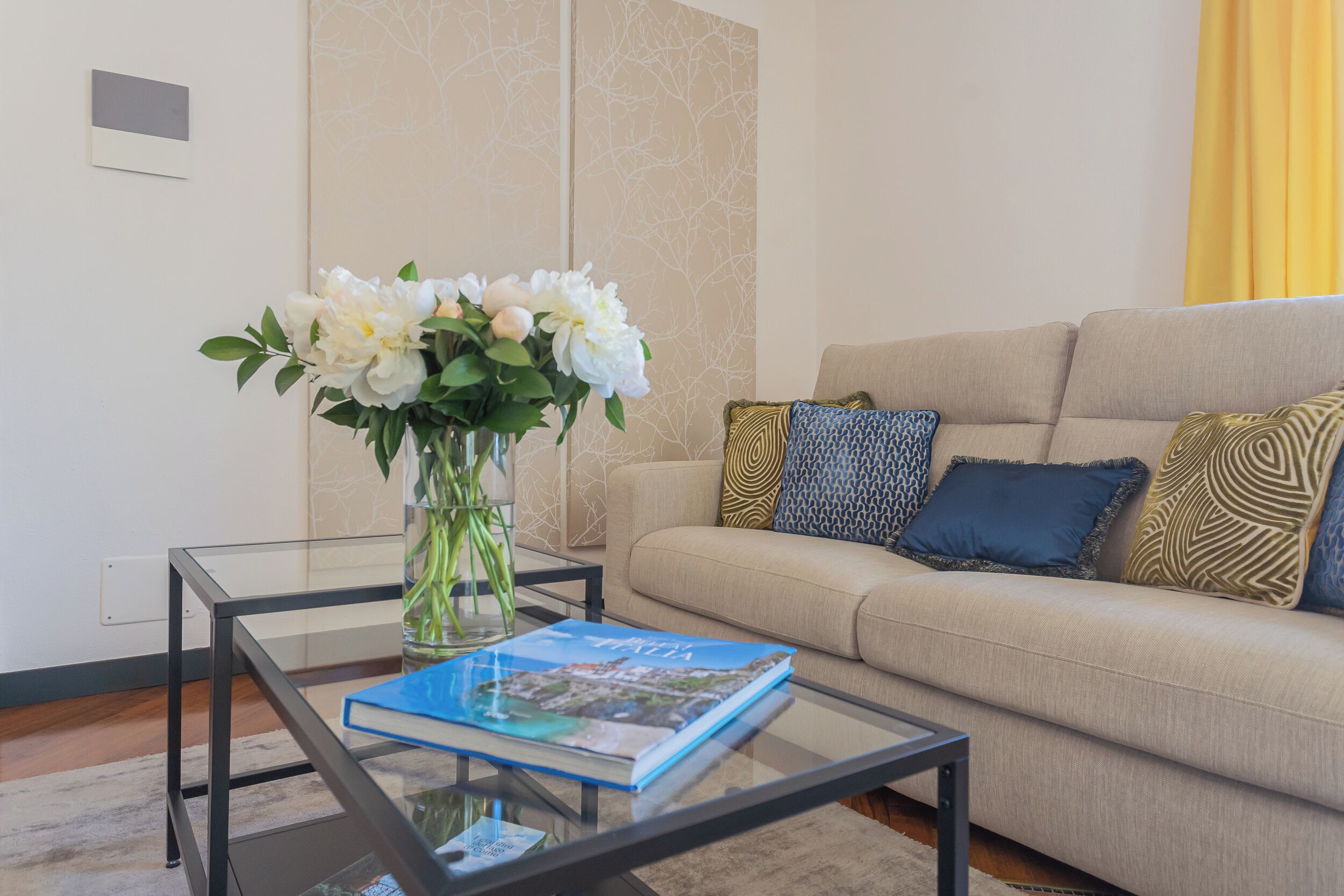 A living room with a glass coffee table, a beige sofa with patterned cushions, and a vase with white flowers.
