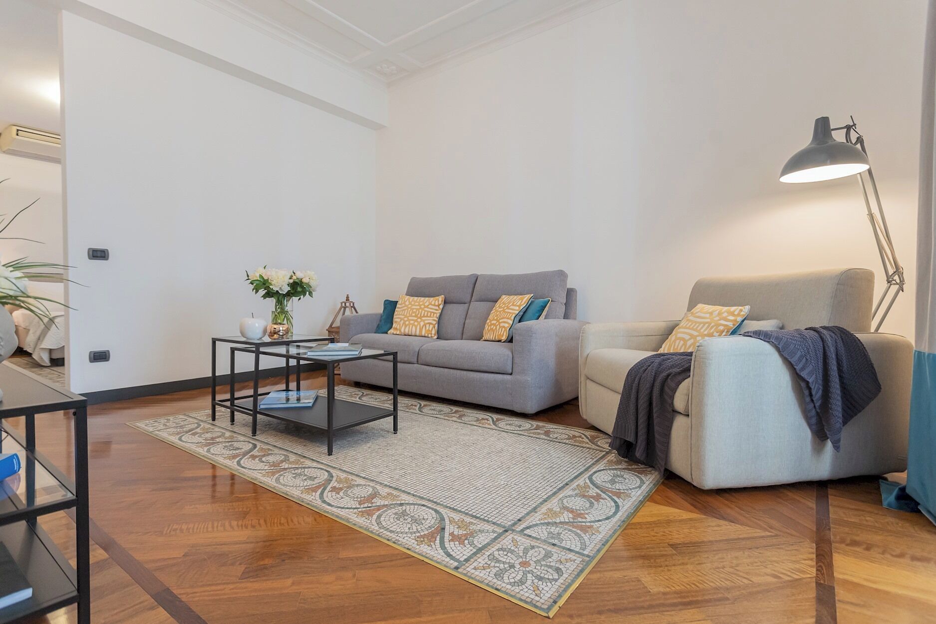 Ein modernes Wohnzimmer mit grauem Sofa, einem beigen Sessel, einem Glastisch und einer Stehlampe.