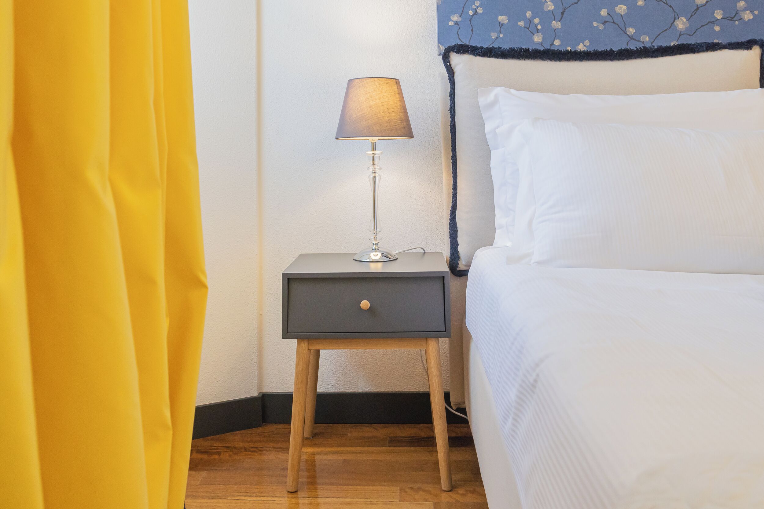 Cama bem arrumada com lençóis brancos, mesa de cabeceira com abajur e cortina amarela semiaberta.