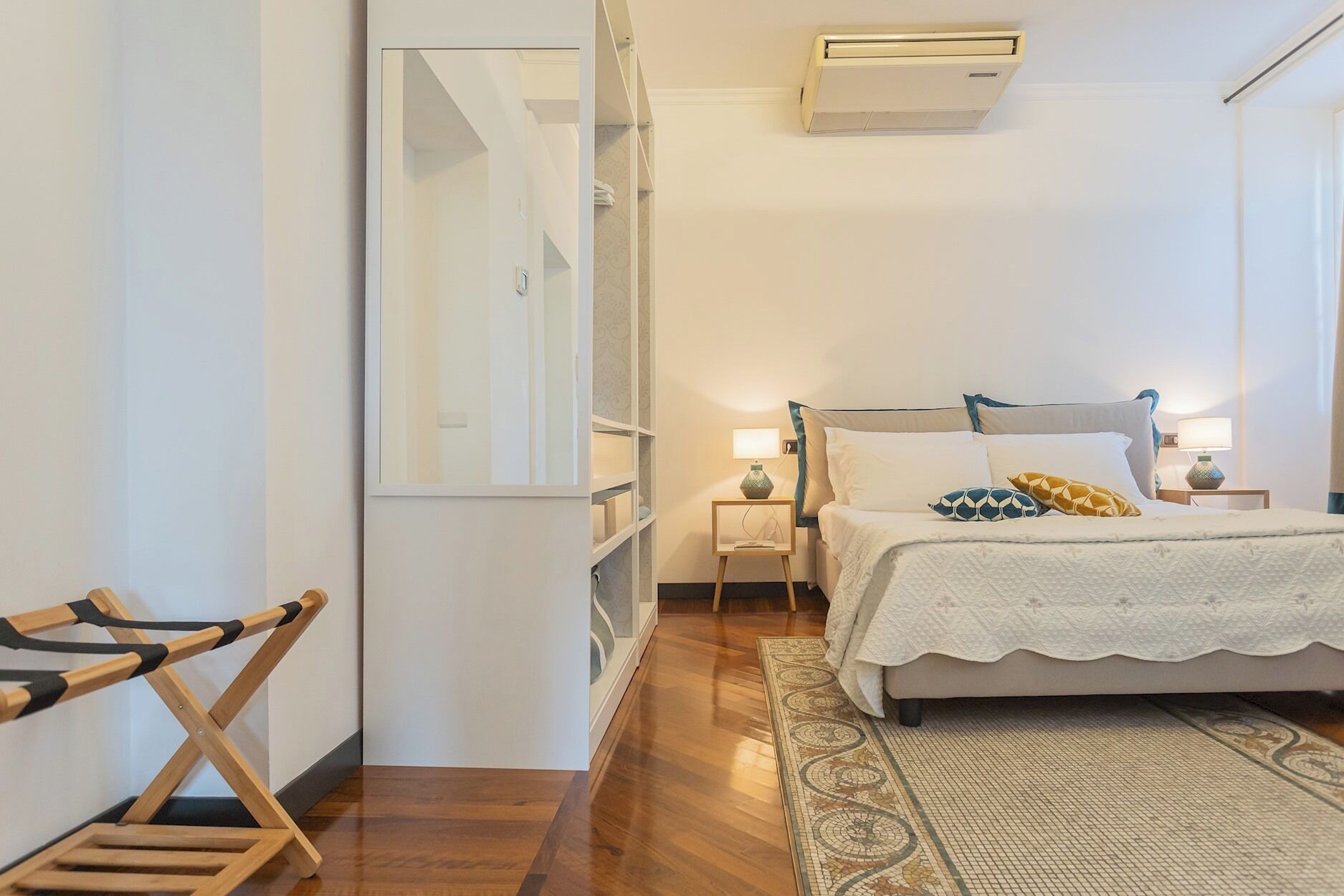 A bedroom with a bed, bedside lamps, a mirror, a wooden chair, and a patterned rug.