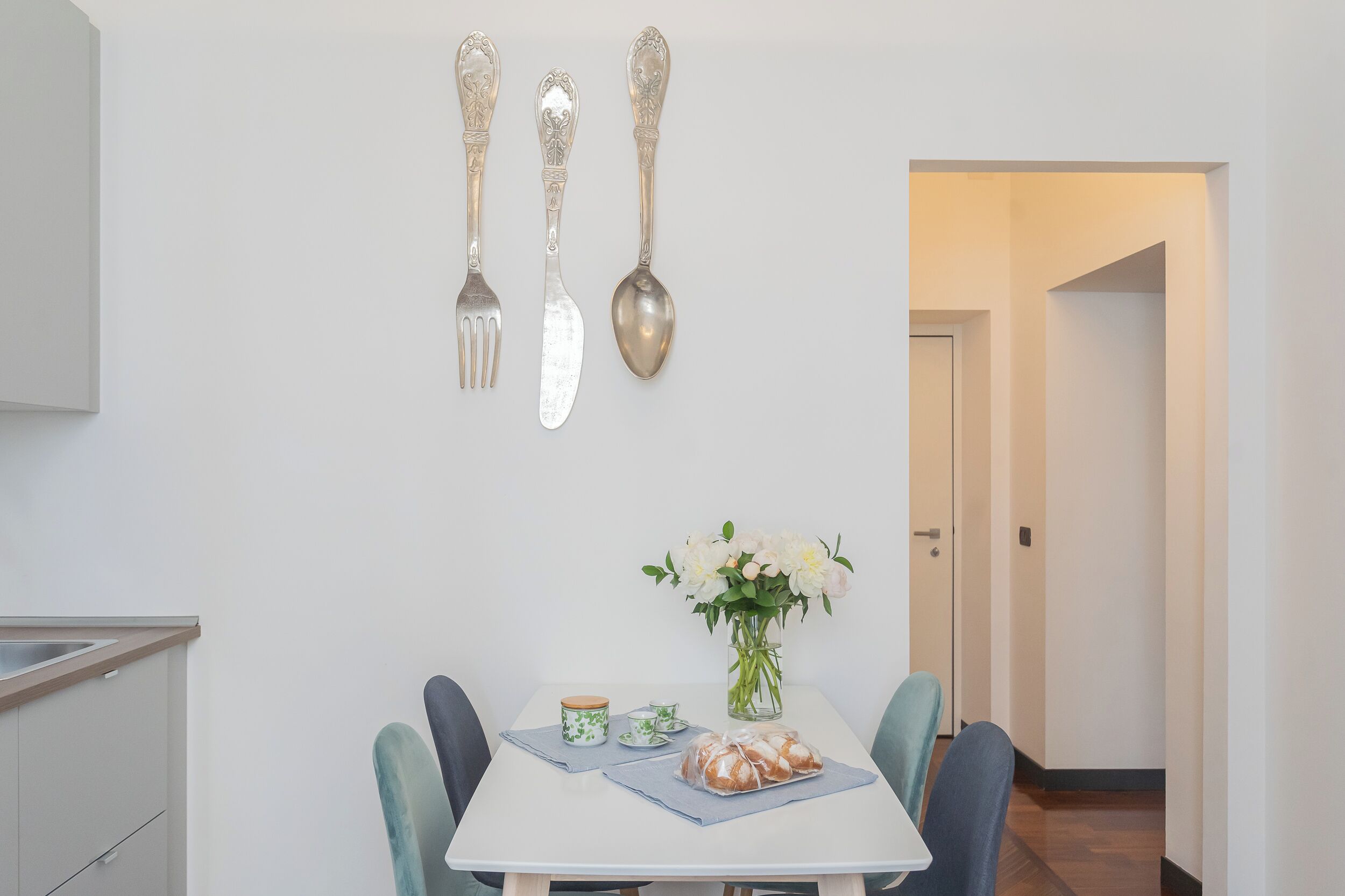 Un comedor con una mesa blanca, dos sillas, un jarrón con flores, un tarro y un pan.