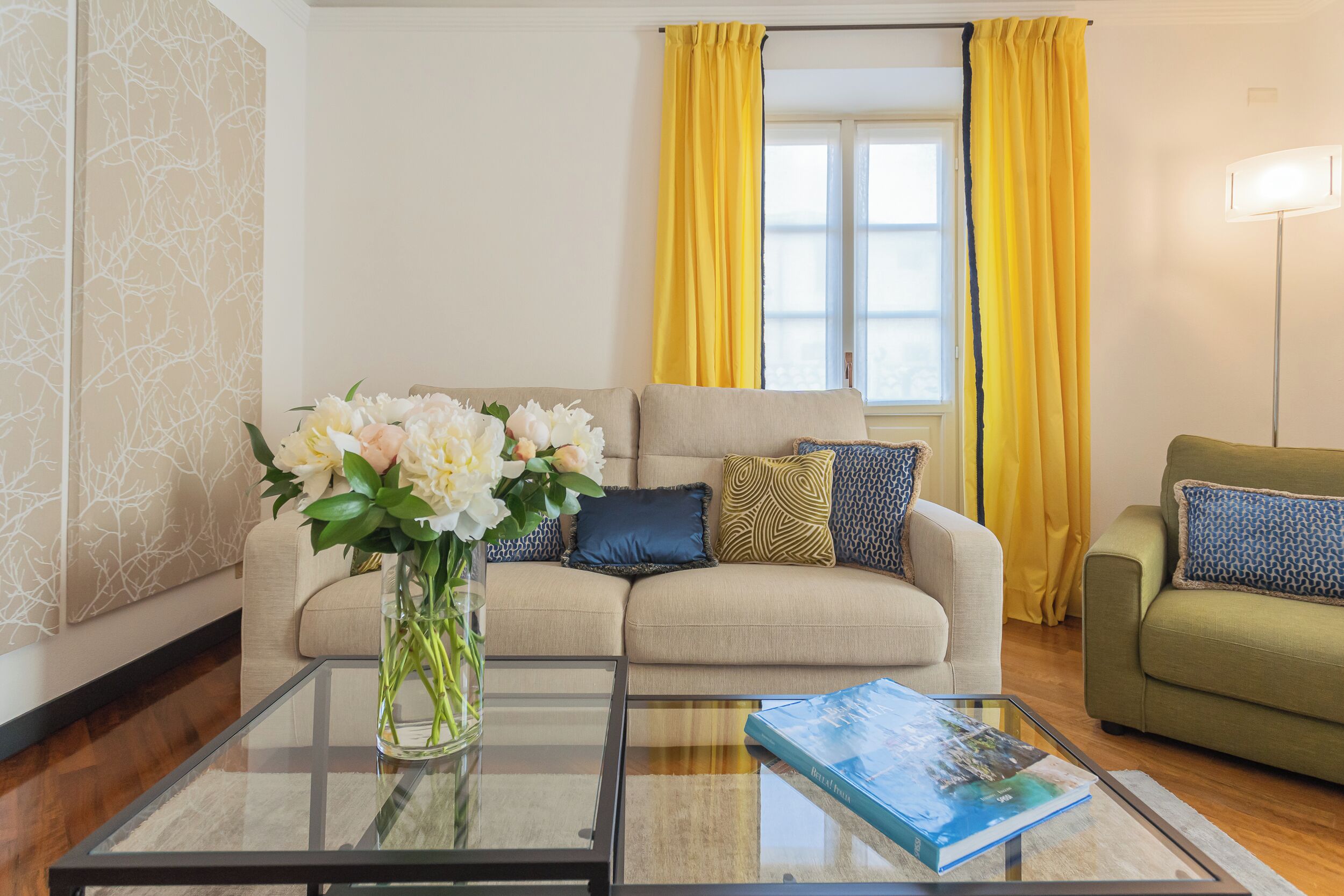 A living room with a beige sofa, yellow curtains, a glass coffee table with a book, and a vase of flowers.