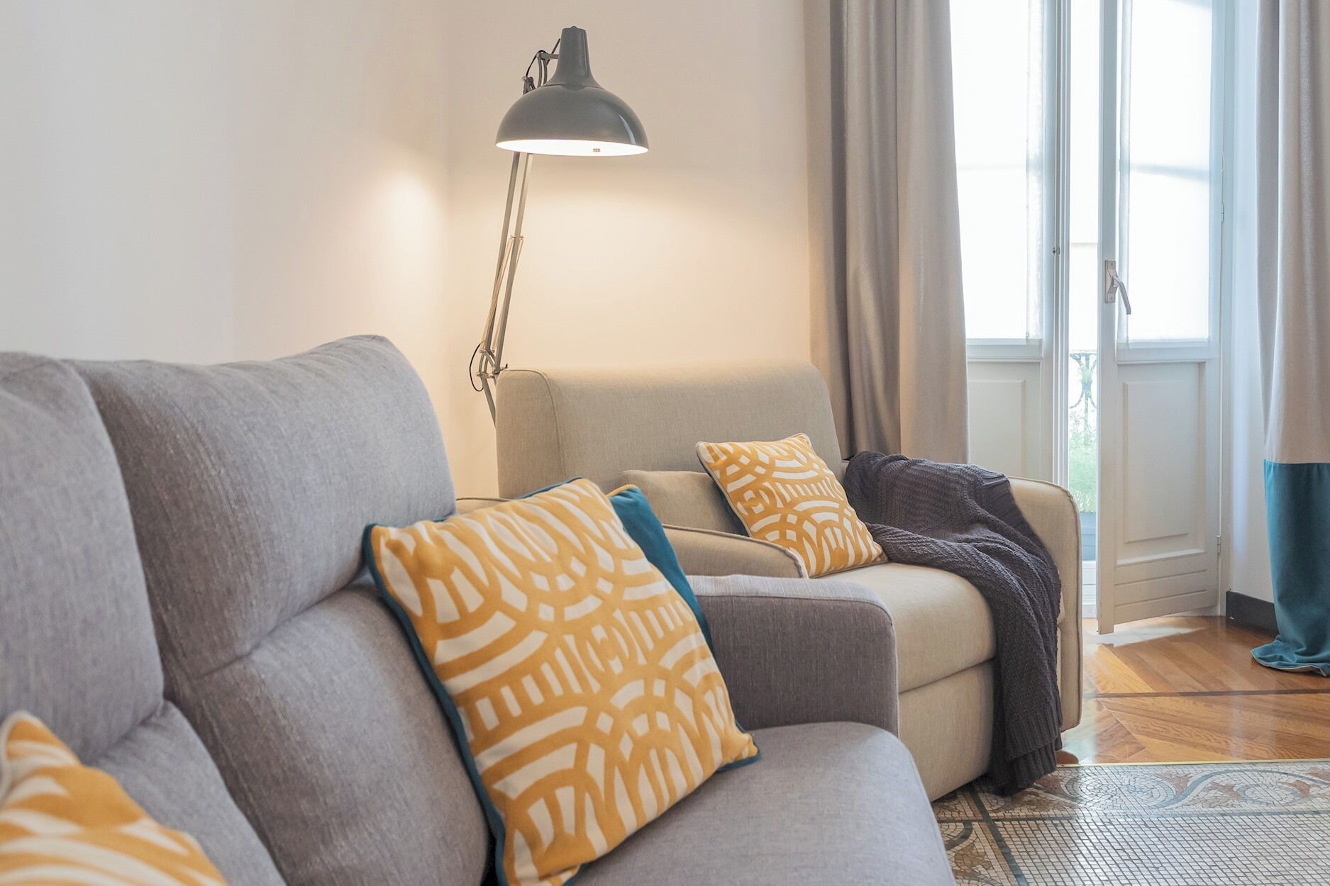 A living room with a grey sofa, patterned cushions, a floor lamp, and a view of the outdoors through a window.