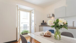 Un coin repas avec une table blanche, des chaises et un vase de fleurs. On aperçoit, au fond, une cuisine équipée d’un four à micro-ondes et d’un évier.