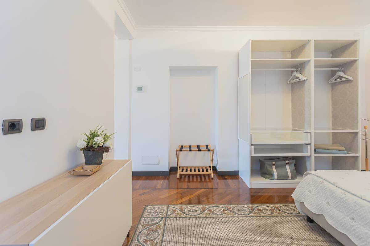Un dormitorio con suelo de madera, una cama con colcha blanca, un armario con espacio para colgar ropa y un pequeño banco de madera.