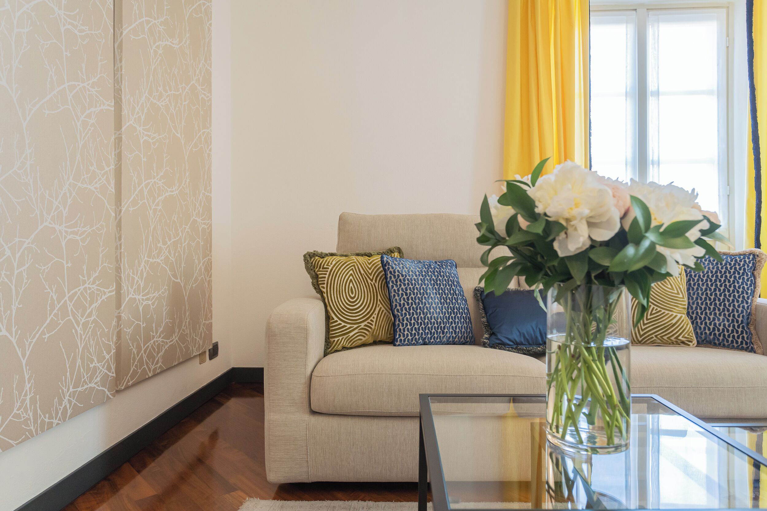 Un salon avec un canapé, des coussins à motifs, une table basse en verre et un vase rempli de fleurs.