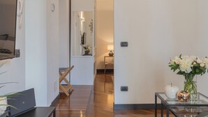 Un hall d’hôtel moderne avec un plancher en bois, une table en verre ornée d’un vase de fleurs, un bureau avec un ordinateur portable et un miroir qui reflète la pièce.