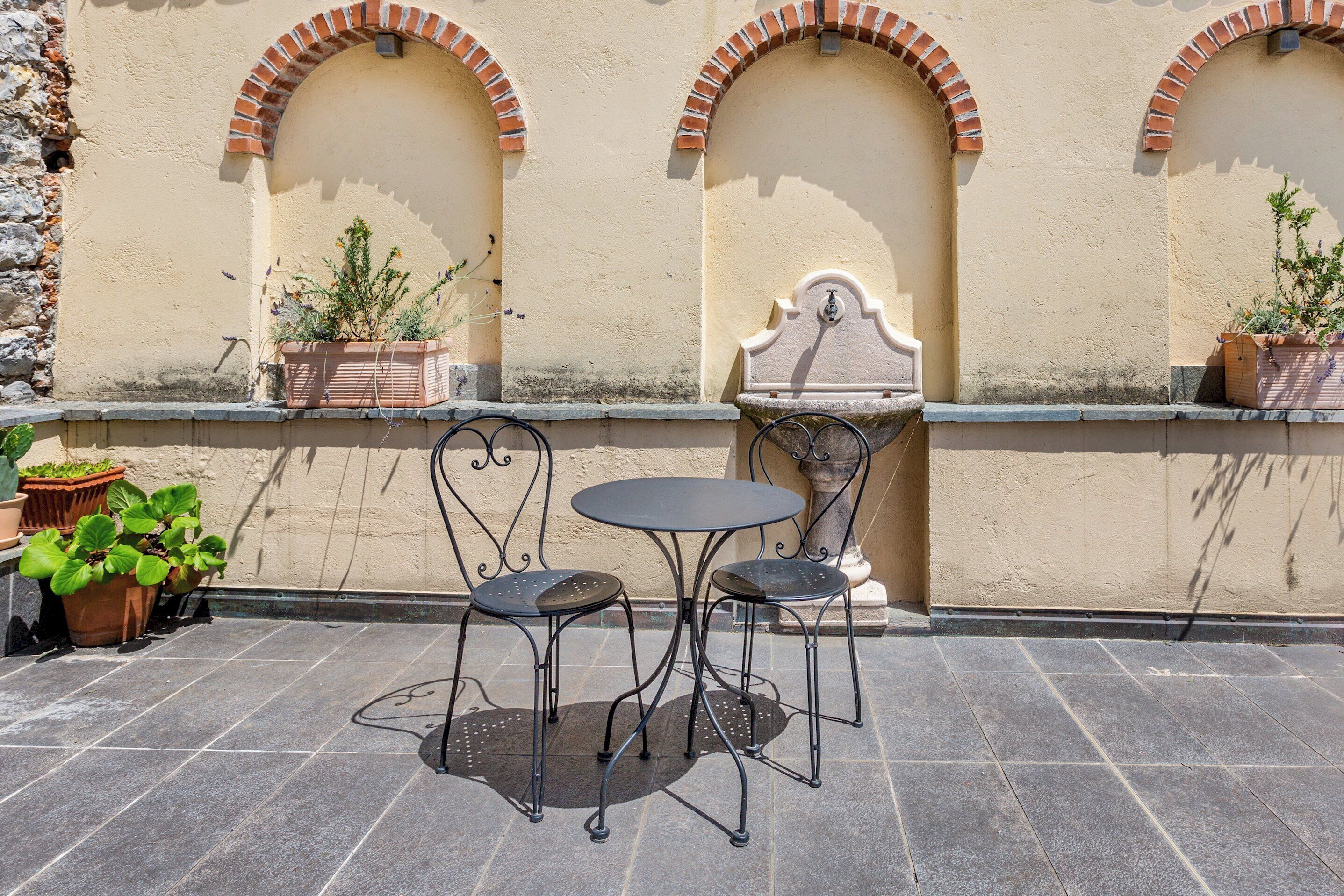 Eine Terrasse mit rundem Tisch, zwei Stühlen, Topfpflanzen und einer Wand mit Rundbögen.
