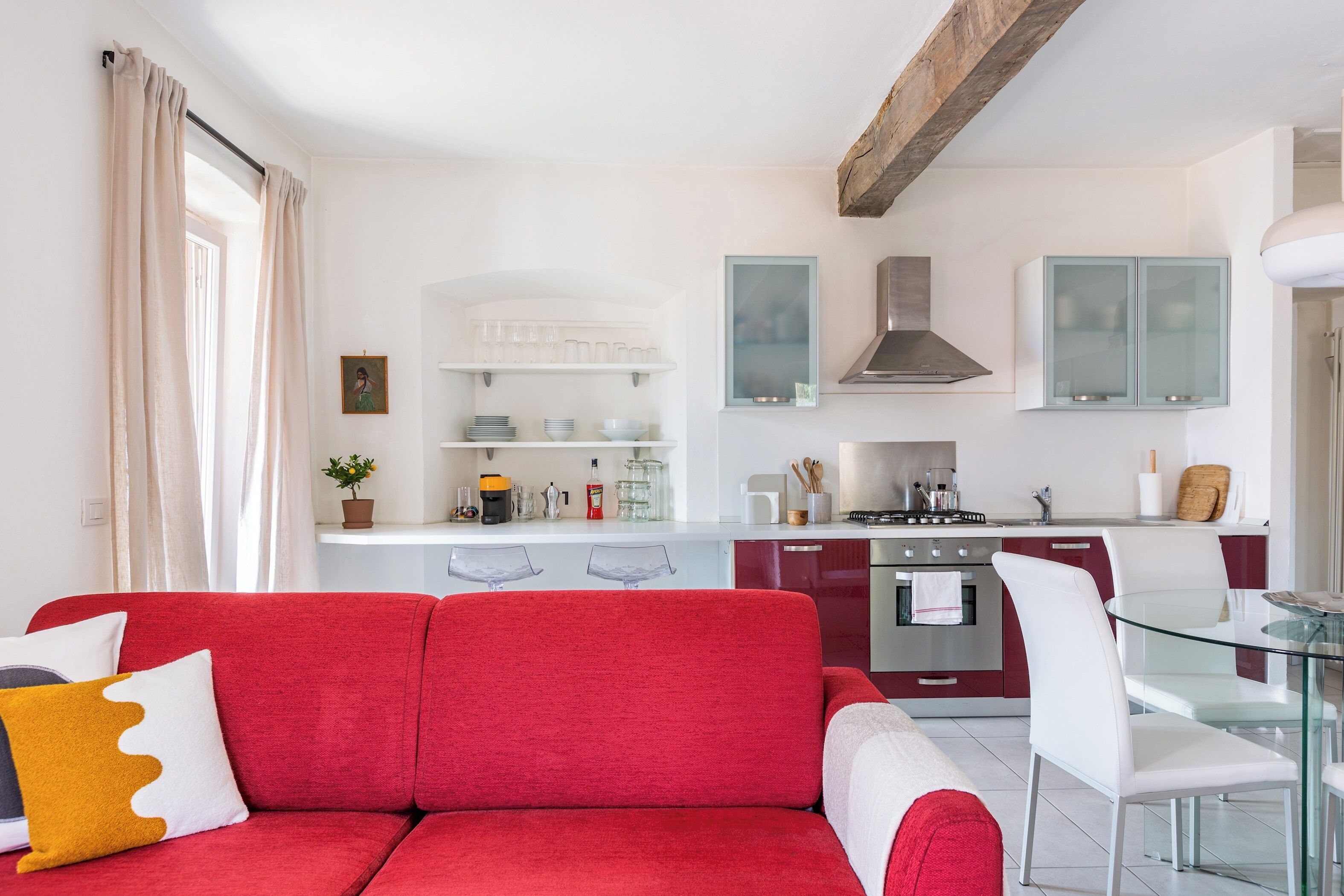 Uma cozinha moderna com armários brancos, uma mesa de jantar de vidro e um sofá vermelho com almofadas decorativas.