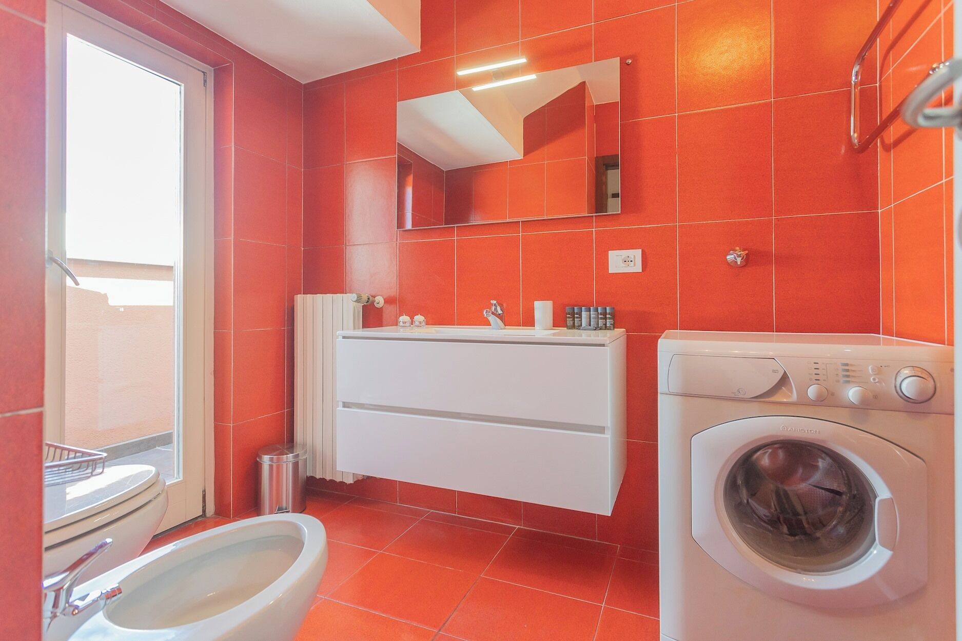 A bathroom with red tiles, a white sink, a mirror, a toilet, and a washing machine.