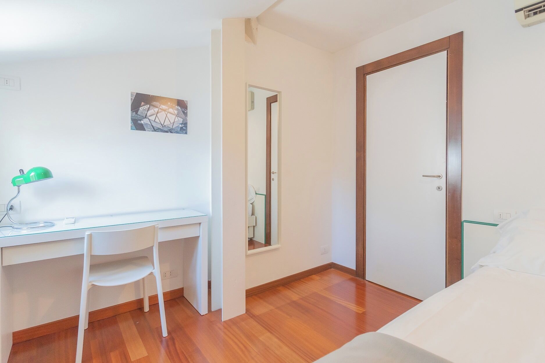 A small, bright room with a white desk, a chair, and a bed.