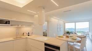 Cocina moderna con armarios blancos, microondas integrado y lavavajillas. Hay un comedor con mesa y sillas, y un amplio ventanal con vistas al mar.