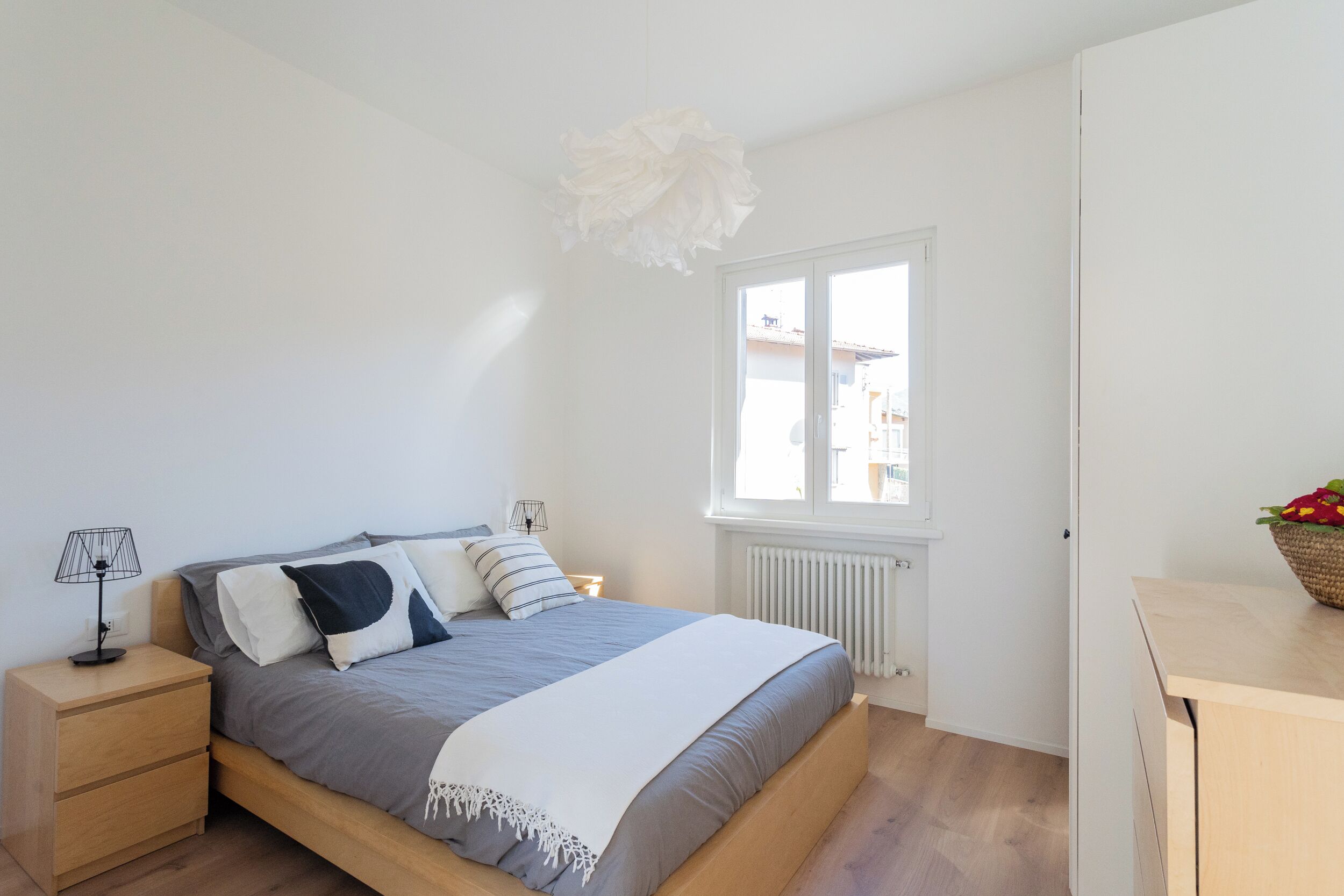 Une chambre à coucher comprenant un lit, une table de chevet, une lampe, une fenêtre, un radiateur et une vue sur les bâtiments environnants.