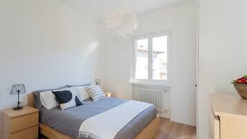 Une chambre à coucher comprenant un lit, une table de chevet, une lampe, une fenêtre, un radiateur et une vue sur les bâtiments environnants.