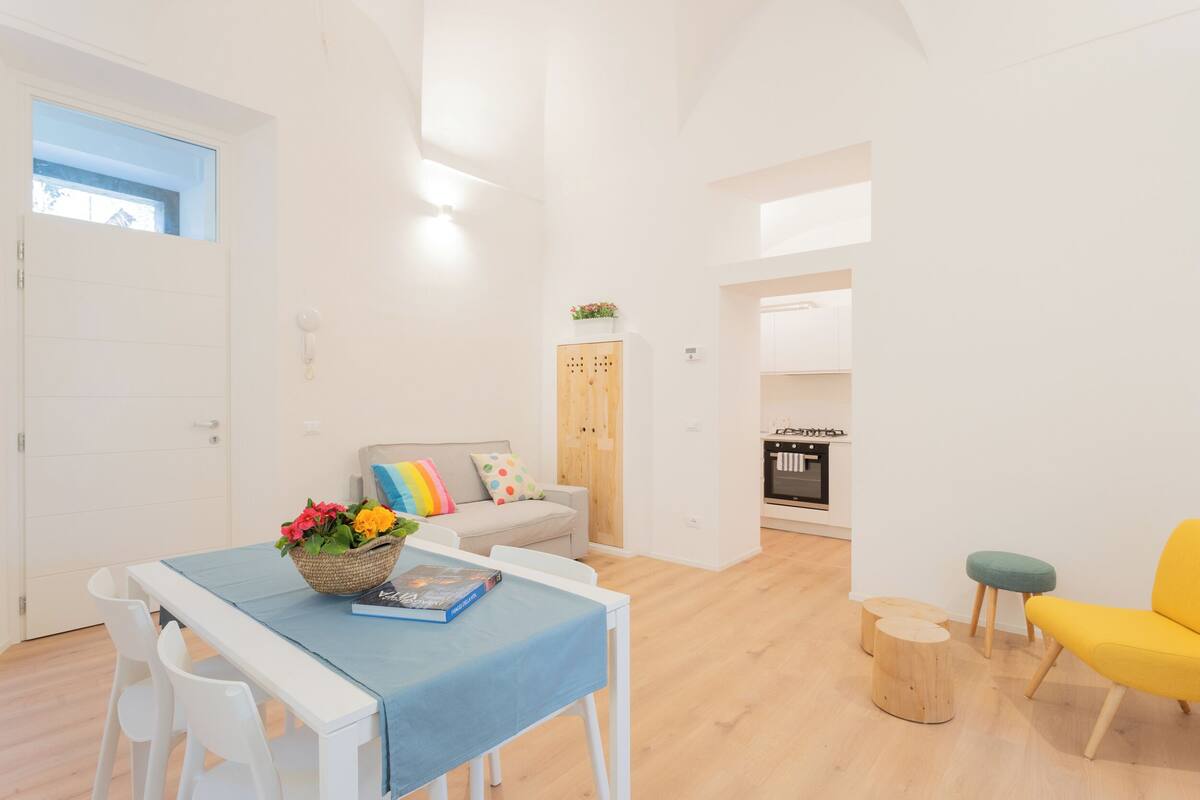 Un salon lumineux et moderne, avec un canapé blanc, un fauteuil jaune et une table à manger recouverte d’une nappe bleue.