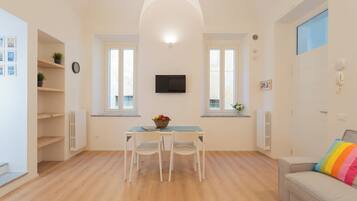 A modern living room with a dining area, a flat-screen TV mounted on the wall, and a window with a view.