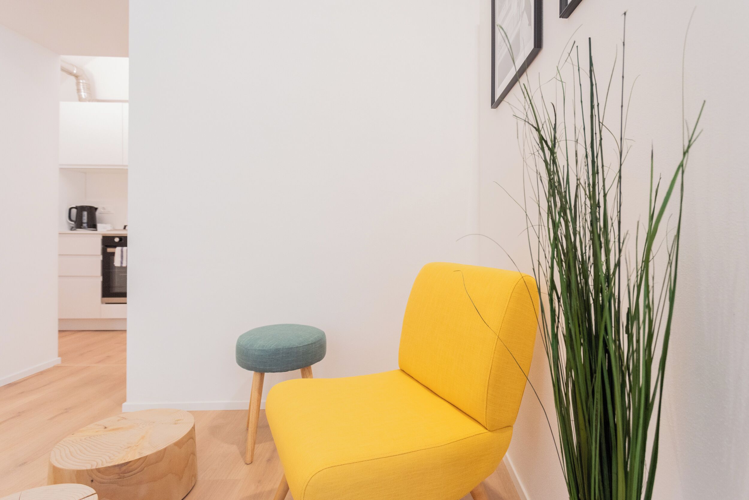 A bright yellow armchair with wooden legs, a small teal stool, and a tall green plant in a modern, minimalistic room with white walls and a wooden floor.