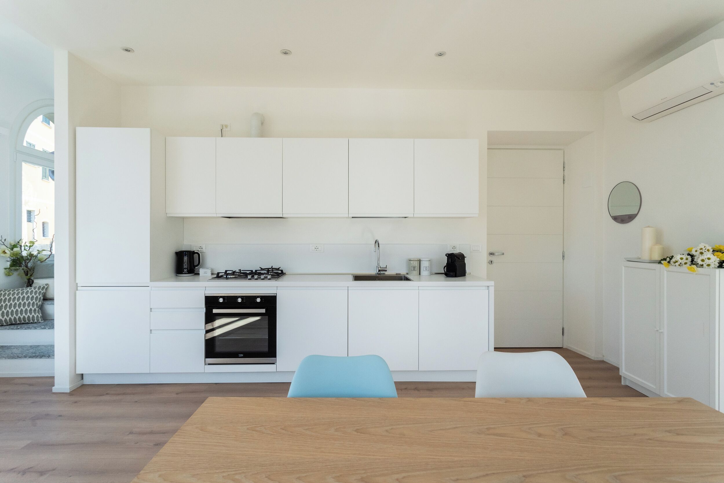 A modern kitchen with white cabinetry, a built-in oven, and a dining area with a wooden table and blue chairs.