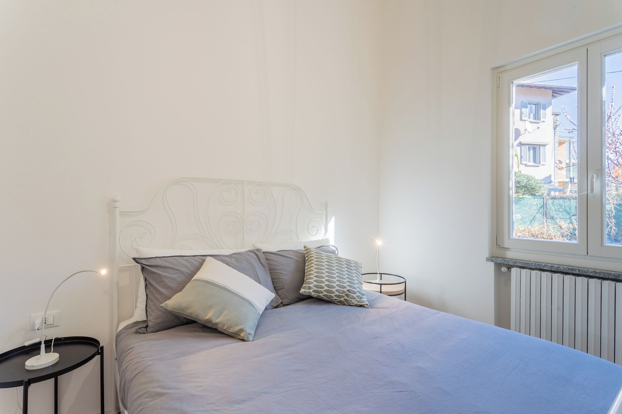 Et soveværelse med seng, puder, et natbord, en lampe og et vindue med udsigt til bygninger.