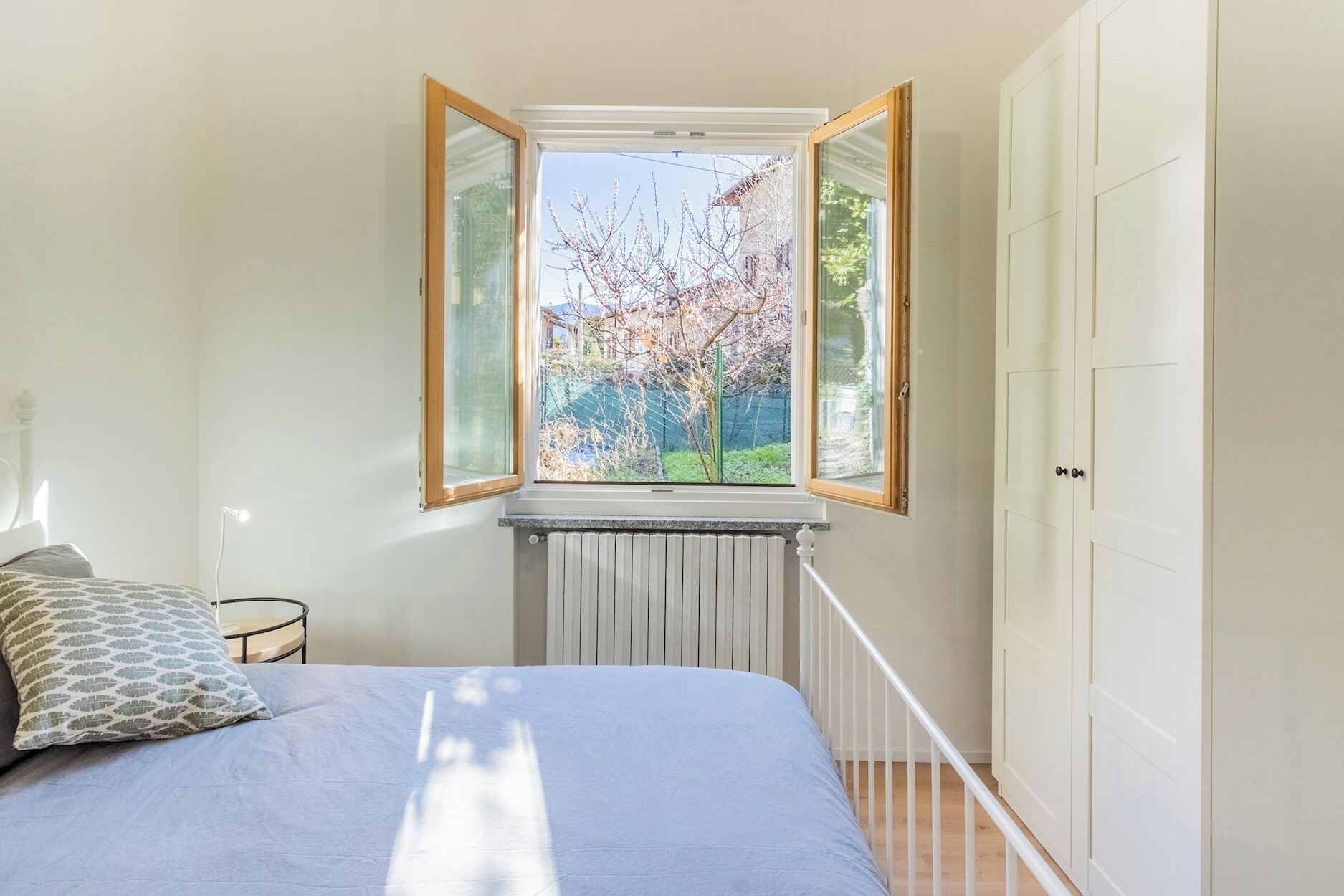 A bedroom with a bed, a nightstand, a radiator, and a window with open shutters.