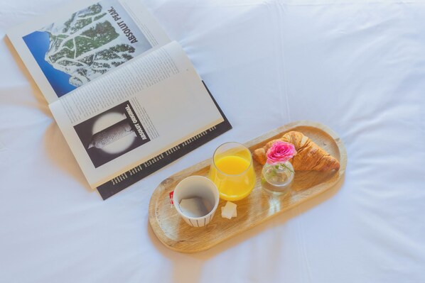 Un livre ouvert sur un lit, à côté d’un plateau contenant un croissant, un verre de jus d’orange, une tasse et un petit vase avec une fleur.