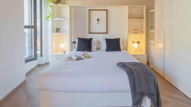 A neatly arranged bedroom with a large bed, bedside tables, a framed picture on the wall, and a window with a plant.