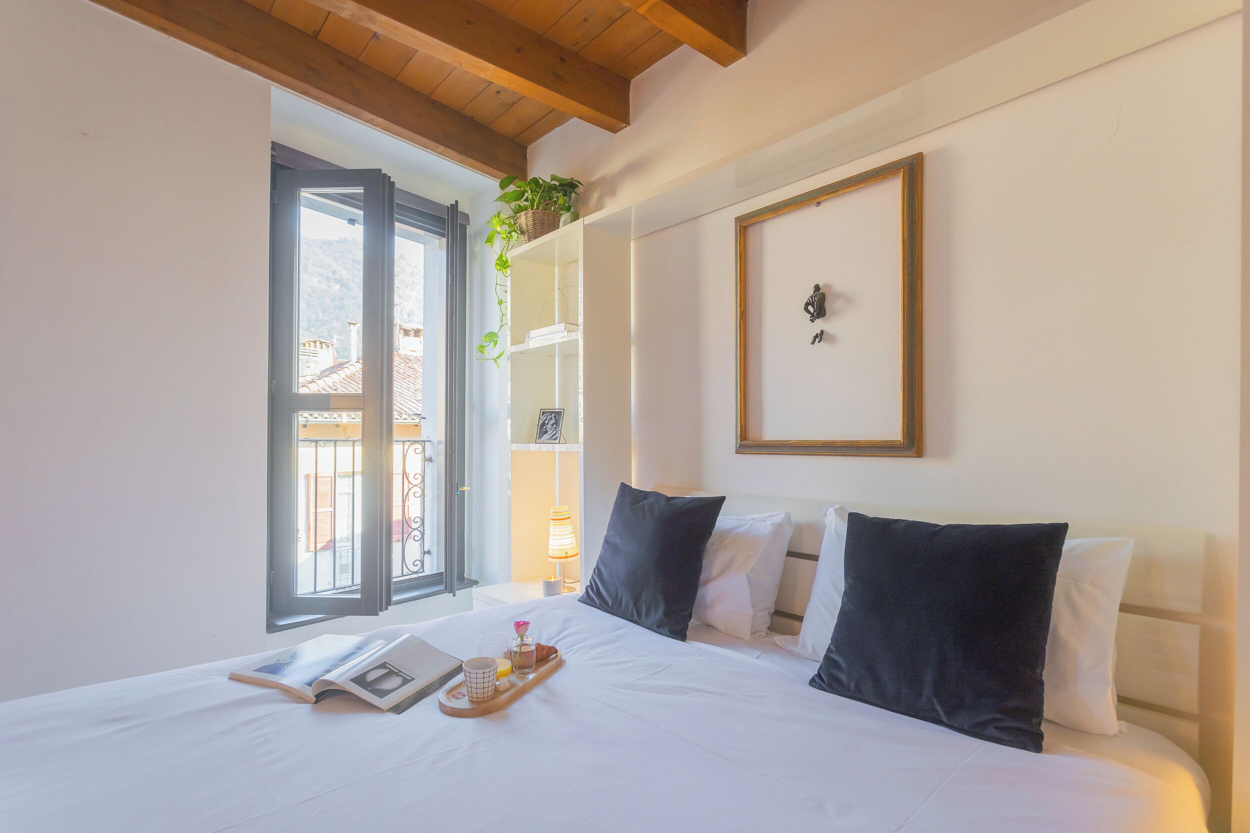 A neatly made bed with pillows, a bedside table with a tray of items, a window with a view of buildings, and a wall-mounted shelf with a plant.