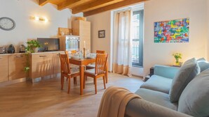 A cozy living room with a wooden ceiling, a dining area with wooden chairs and a table, a kitchen with modern appliances, and a colorful abstract painting on the wall.