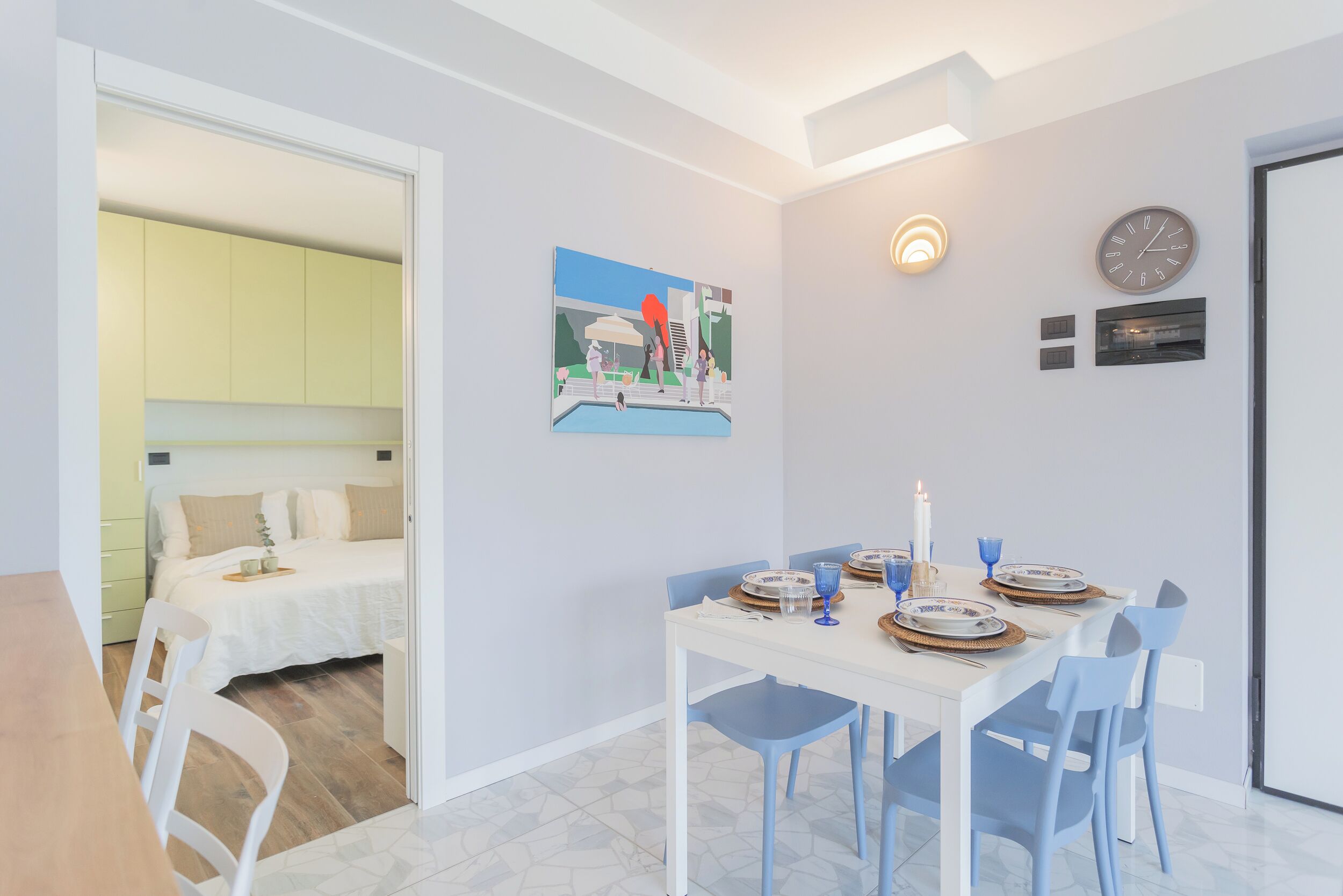 A dining area with a white table set for four, blue chairs, and a painting on the wall.