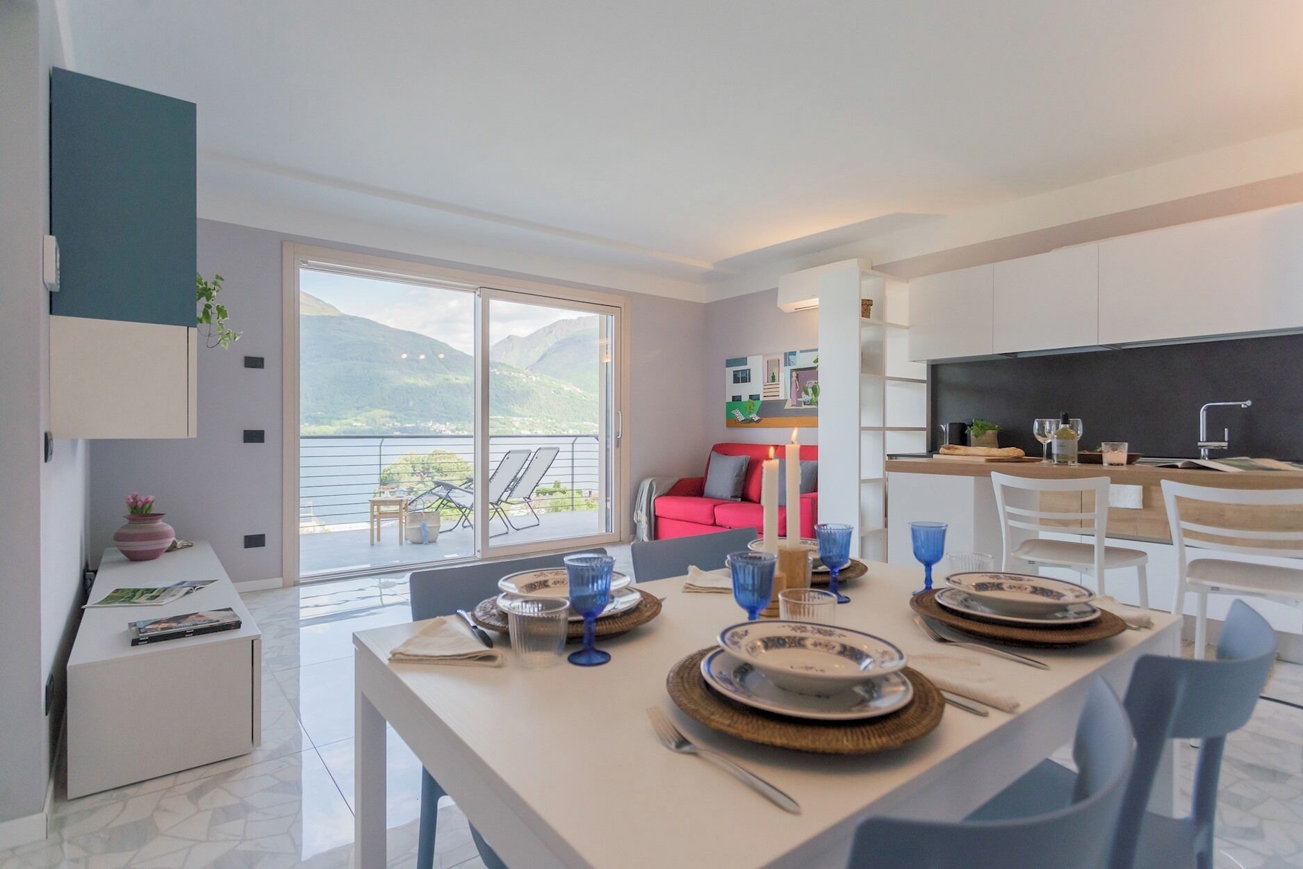 A modern kitchen with a dining area set for a meal, a red sofa, and a balcony with a scenic view.
