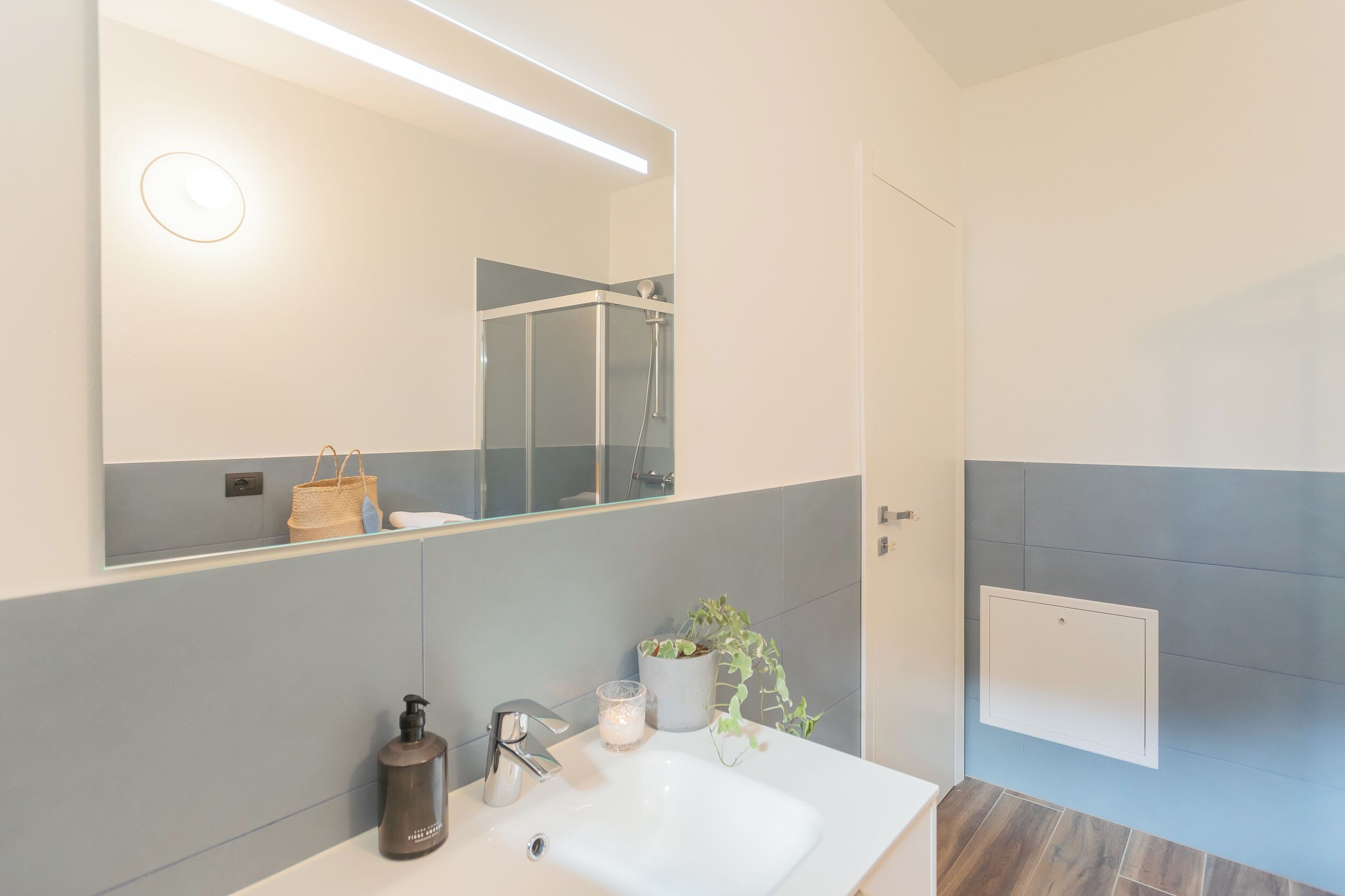 A modern bathroom with a large mirror, a white sink, a shower, and a potted plant.