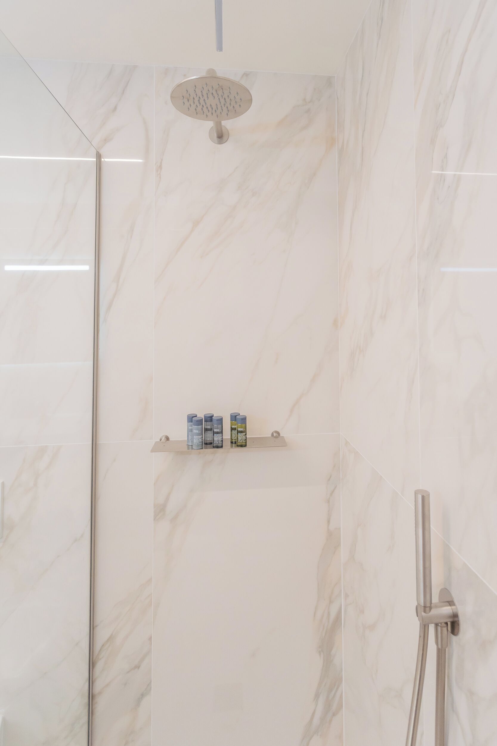 A modern shower area with a glass door, marble walls, and a shelf with toiletries.