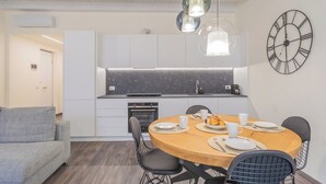 Una cocina moderna con armarios blancos, una encimera negra y una mesa redonda con sillas. Hay un reloj en la pared y una lámpara colgante sobre la mesa.