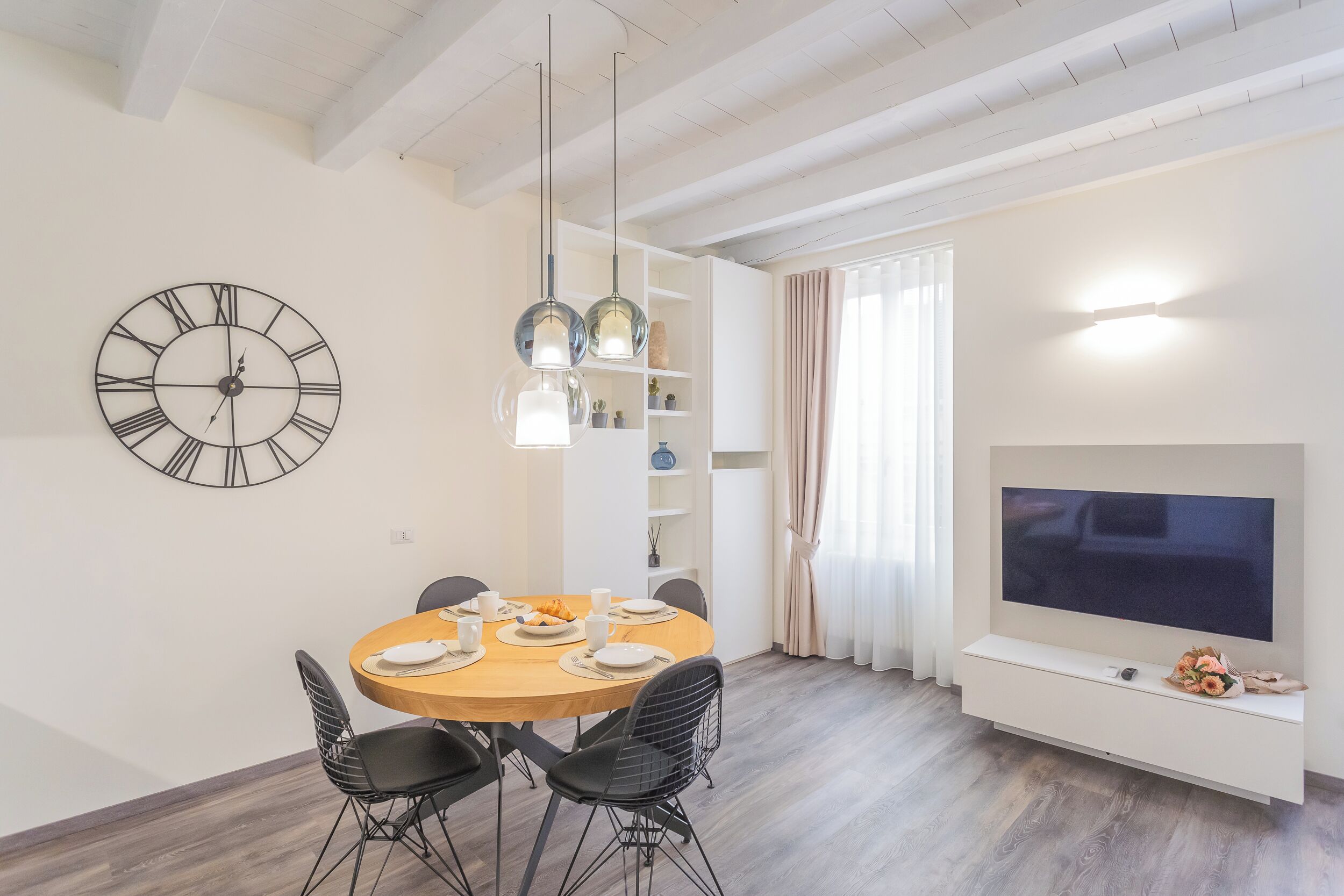 A modern dining area with a round table, chairs, a wall clock, pendant lights, a TV, and a shelf with decorative items.