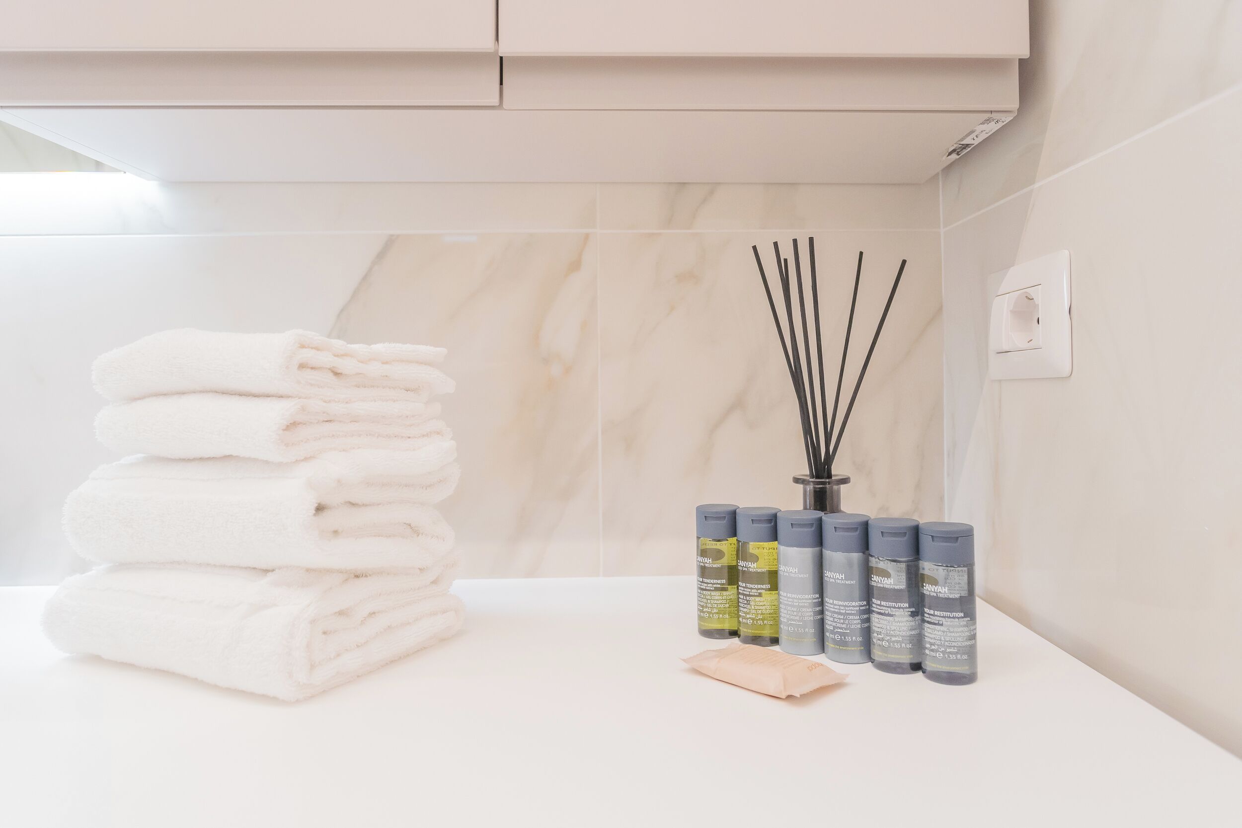 A neatly arranged bathroom with a stack of white towels, a set of reed diffusers, and a soap bar on a white countertop.