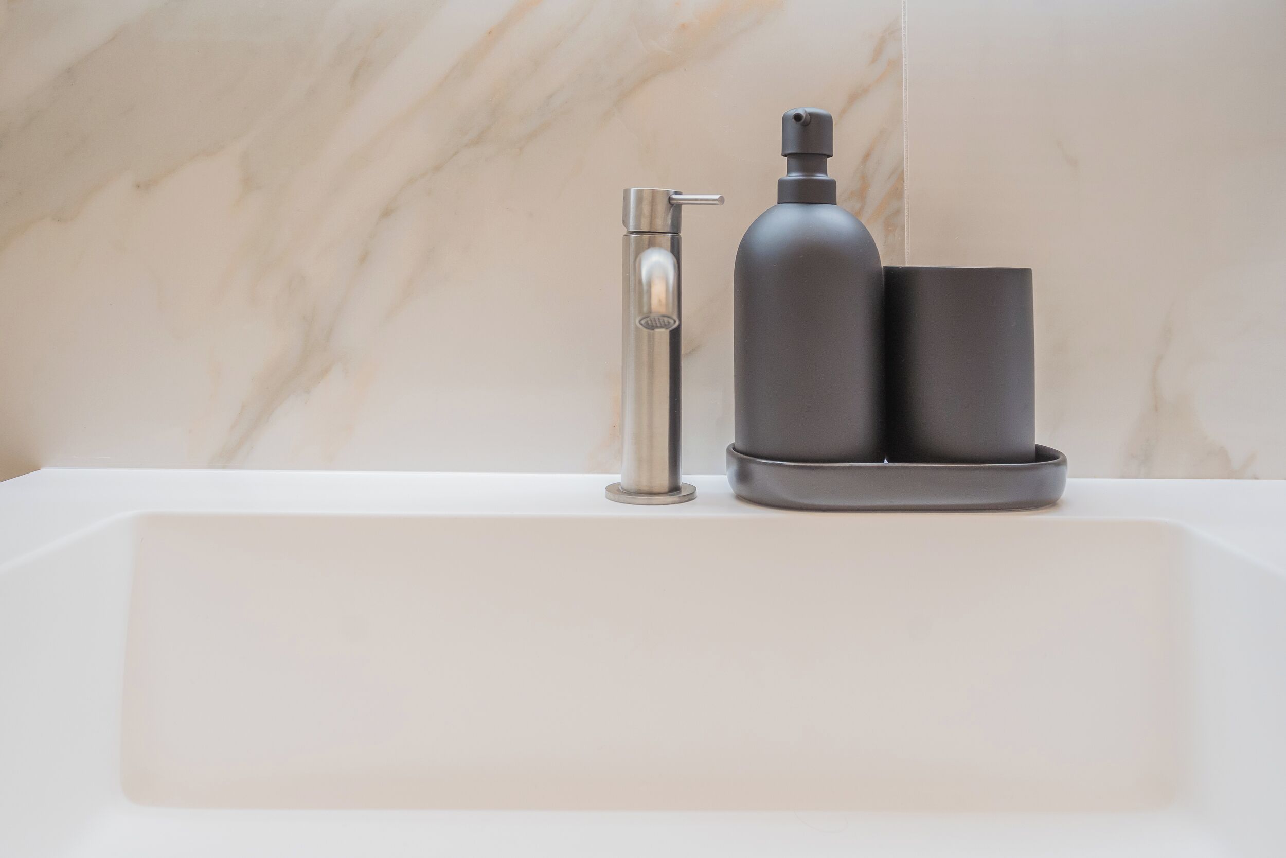 A modern bathroom with a marble wall, a chrome faucet, and a black soap dispenser.