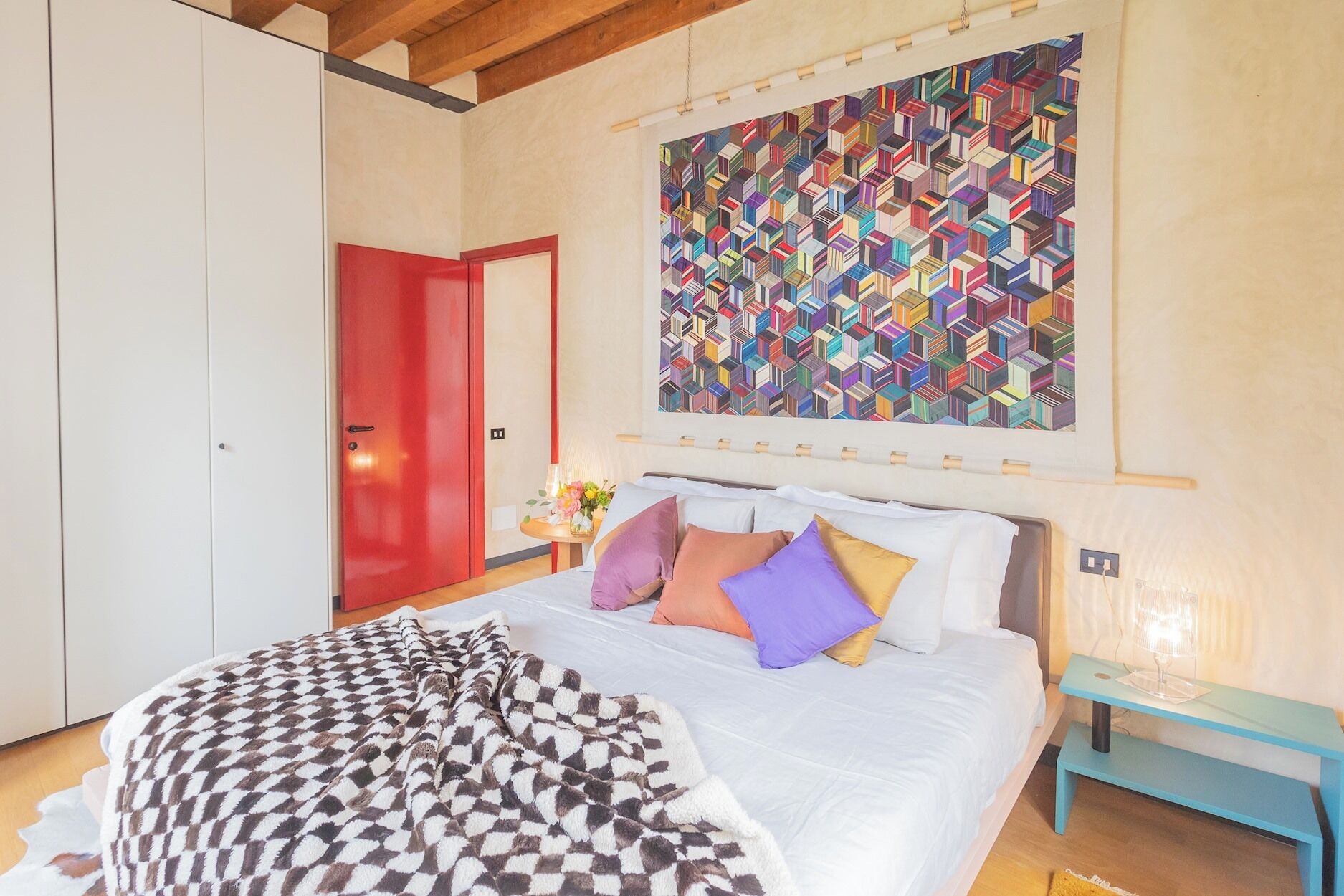 Un dormitorio con cama, almohadas, una colcha de cuadros, una puerta roja y un colorido arte mural geométrico.