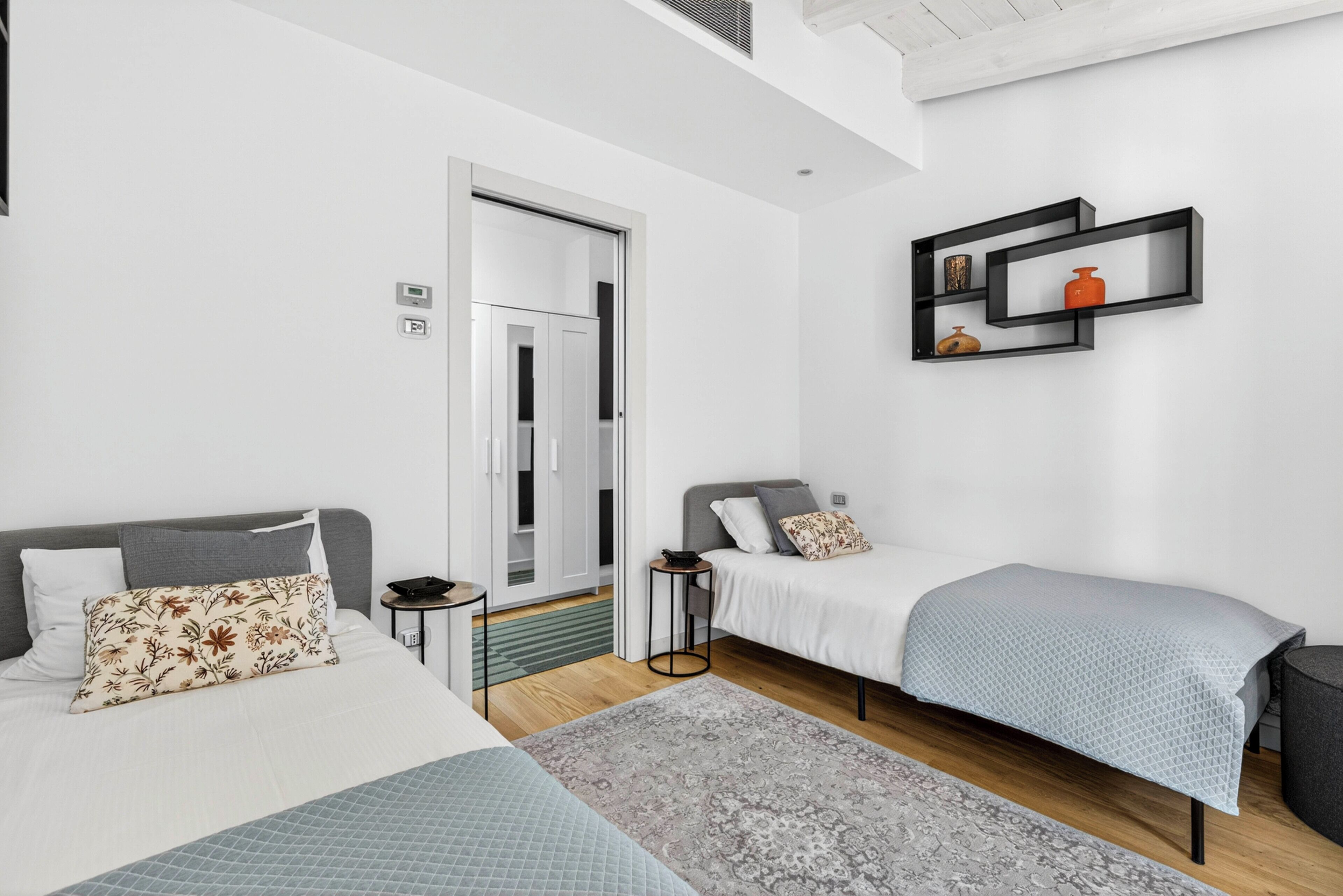 Une chambre moderne avec deux lits, une armoire, une petite table et une étagère murale agrémentée d’objets décoratifs.