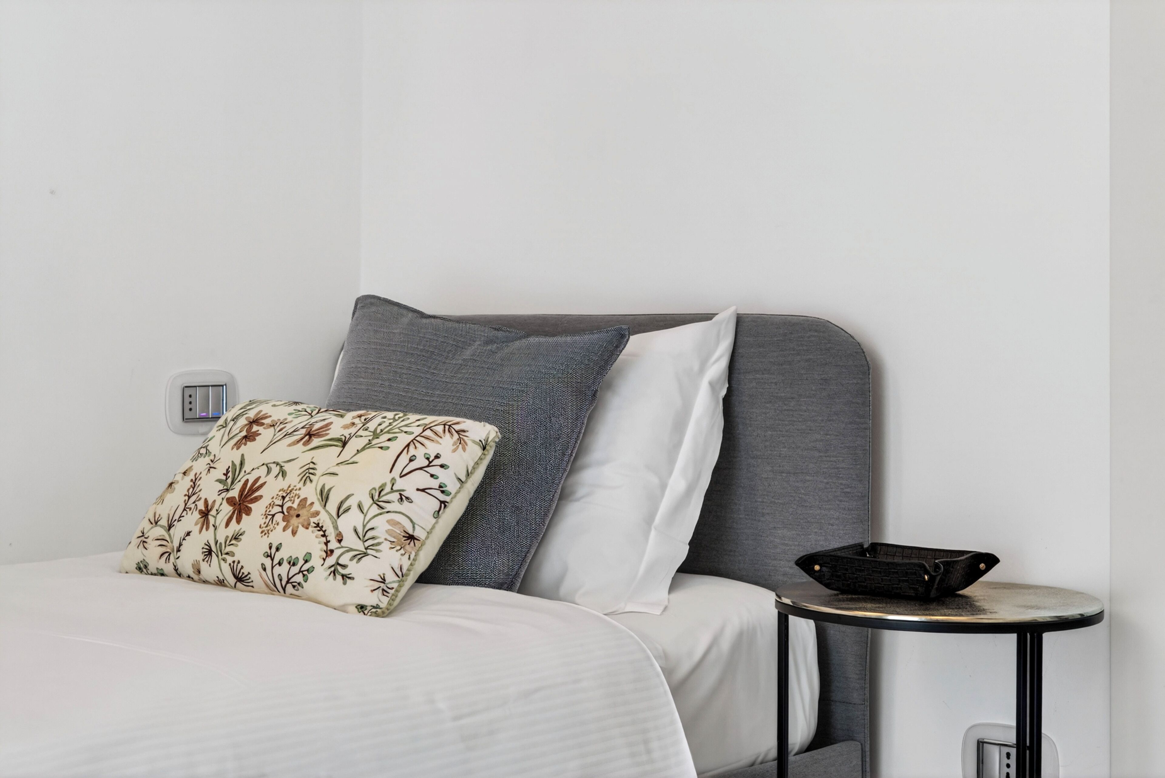 A bed with a floral pillow, a grey headboard, and a small round table with a black tray.