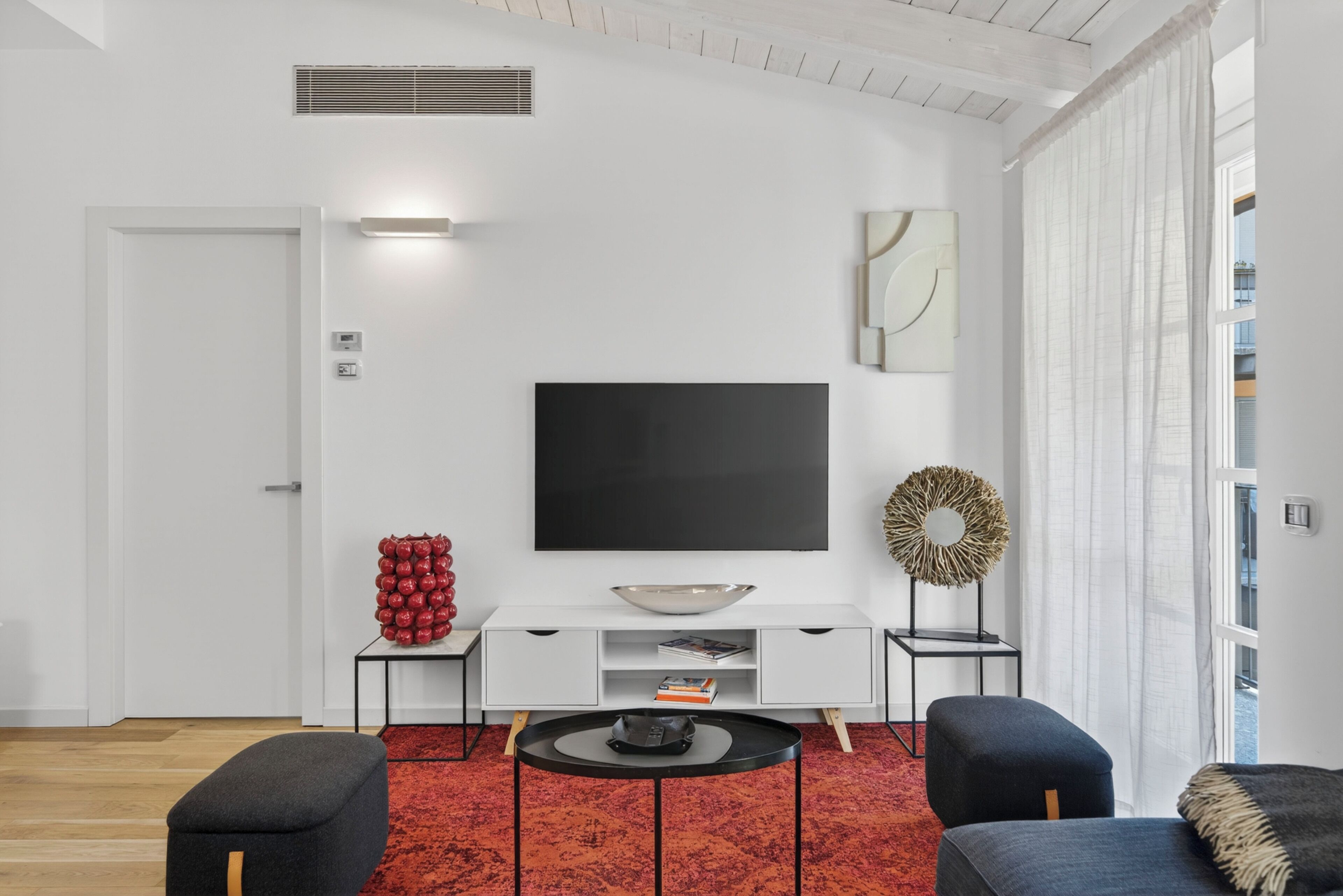 A modern living room with a flat-screen TV, white furniture, a red rug, and a view through a window.