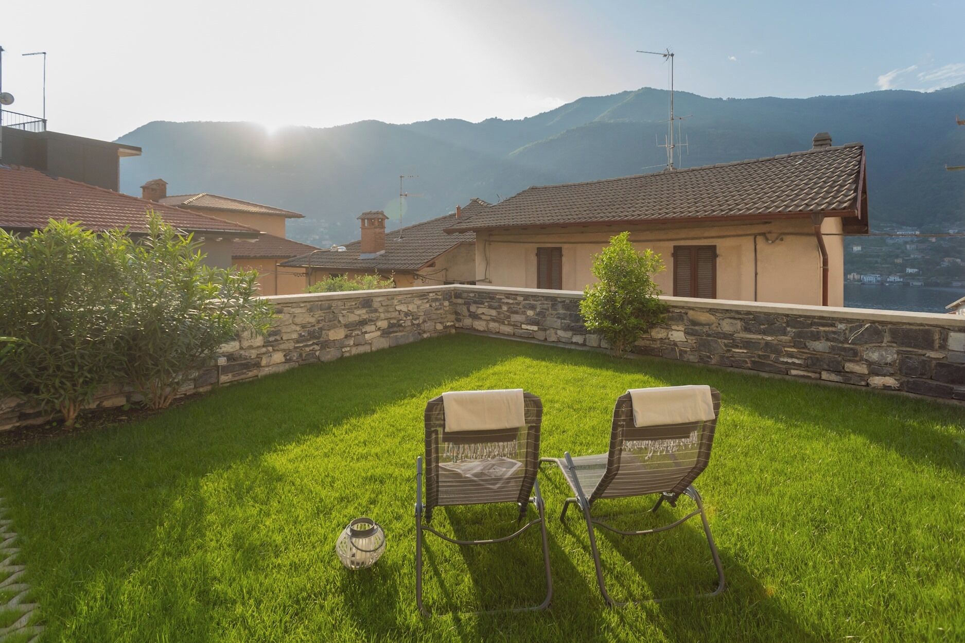 Eine Terrasse mit zwei Stühlen und einer Laterne, umgeben von einer Steinmauer und einem Rasen, mit Blick auf eine gebirgige Landschaft.