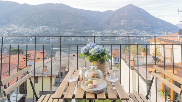 Un balcon avec une table dressée pour le thé, un vase rempli de fleurs et une vue sur un lac et des montagnes.
