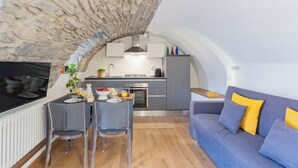 Un espace de vie compact, agrémenté d’une arche en pierre menant à une cuisine, d’un coin repas avec des chaises et d’un canapé bleu avec des coussins jaunes.
