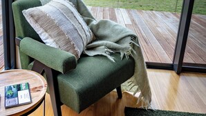Un sillón verde con un plaid, una mesita de madera con un folleto y vista a un lago a través de amplios ventanales.