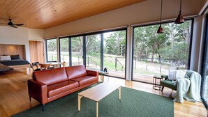 Amplia sala de estar con un sofá de cuero marrón, una mesa de centro de madera y una alfombra verde. Grandes ventanales ofrecen vistas al exterior.