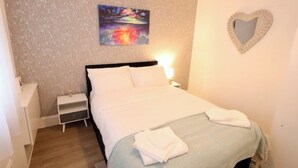 A neatly made bed with white linens, a small bedside table with a lamp, and a wall-mounted heart-shaped mirror. - Modern, Spacious 3 Bedroom home Watford Junction (Watford)