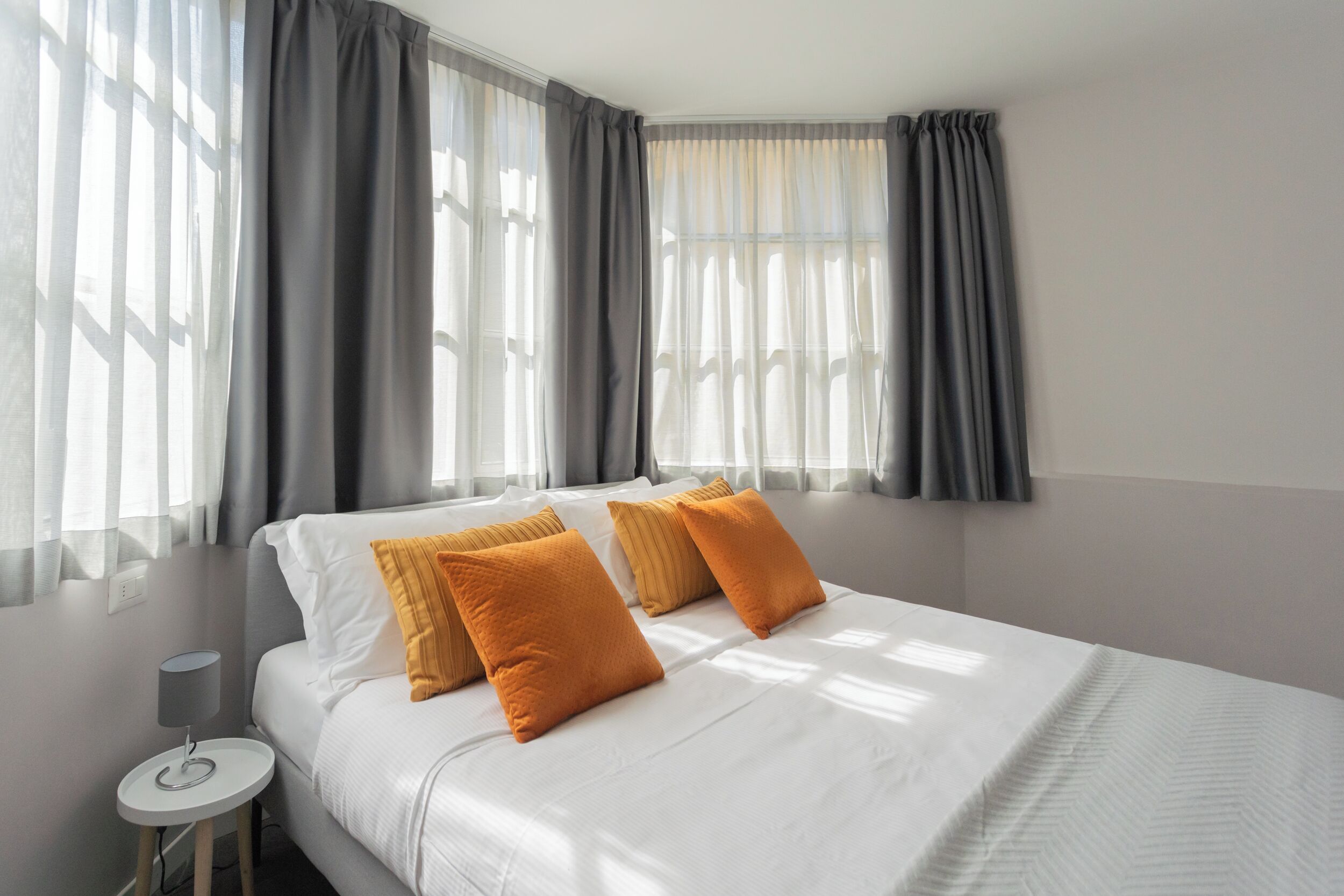 Una cama bien hecha con sábanas blancas y tres almohadas anaranjadas, una mesita de noche con una lámpara y una ventana con cortinas grises.