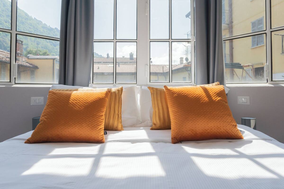 A bed with white linens and two orange pillows, a window with a view of buildings and mountains, and a gray curtain.