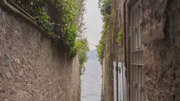 Un estrecho callejón con un sendero adoquinado que lleva a una masa de agua, flanqueado por muros de piedra y una verja de metal.