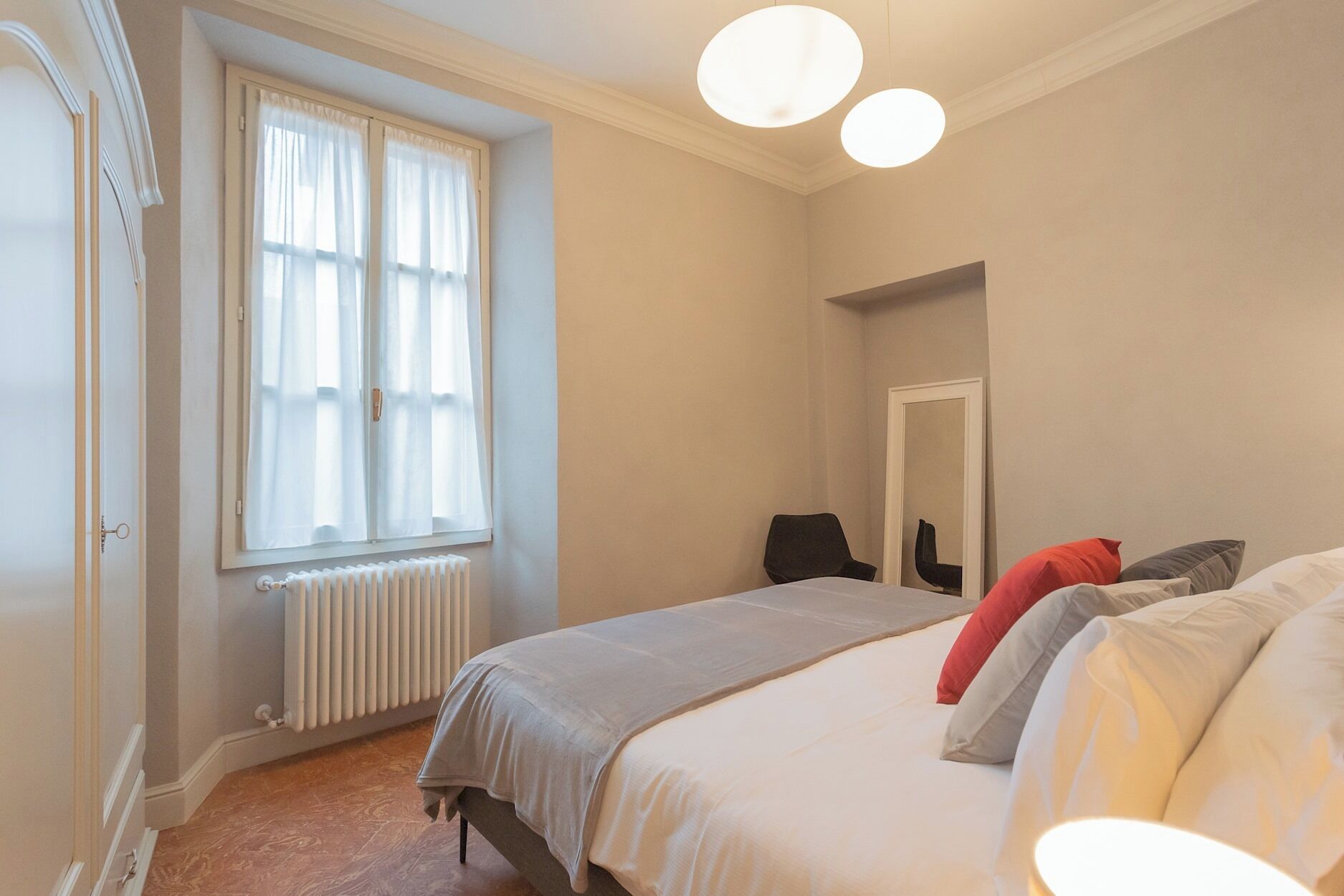 Ein Schlafzimmer mit einem Bett, einem fensterbelaubten Fenster, einem Heizkörper, einem Stuhl und einem Spiegel an der Wand.